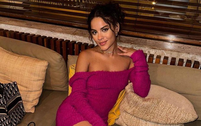 A atriz Bella Campos posa para foto publicada no Instagram, sentada em sofá, com vestido rosa, ombros de fora, expressão séria