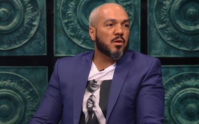 Foto de Belo; ele está sentado, veste camiseta branca e terno azul