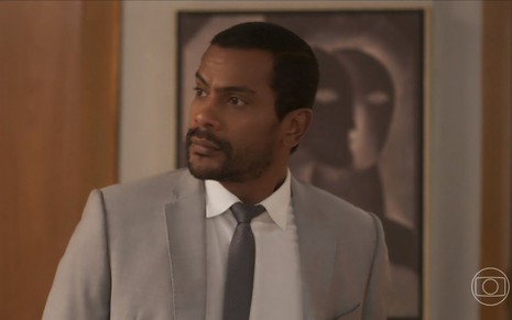 Em cena de Vai na Fé, Samuel de Assis está usando terno cinza e gravata; ele está olhando para alguém