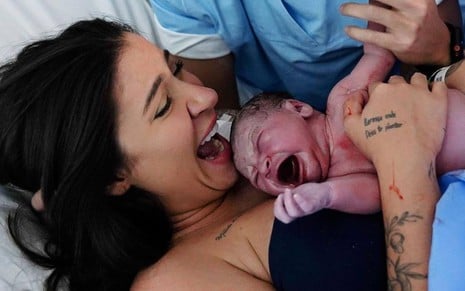 Bianca está deitada, com a boca aberta, ela olha para o filho; o bebê está deitado no peito da influenciadora e chora
