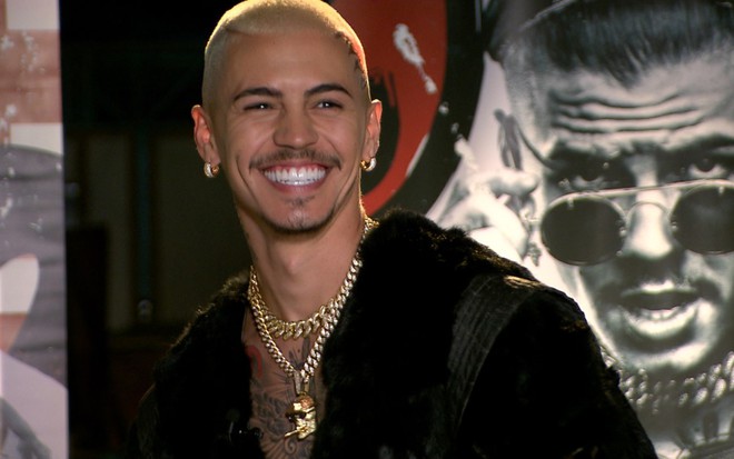 Biel de cabelo loiro, sorrindo, em um cenário que exibe ao fundo uma foto sua em um painel
