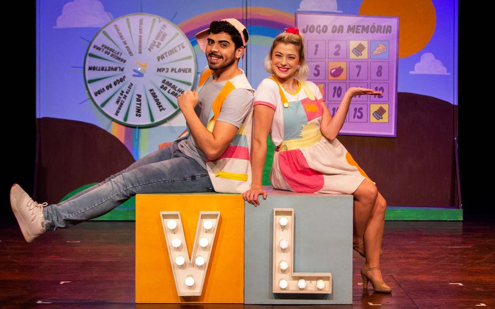 Vitor Rocha e Luiza Porto estão em cenário que simula programa infantil, com direito a roda de prêmios