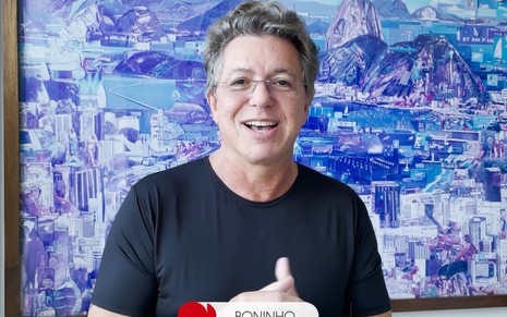 J.B de Oliveira, o Boninho, com uma camisa preta e um fundo azul em sua casa, no Rio de Janeiro