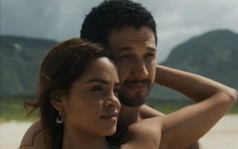 Em cena de Travessia, Romulo Estrela e Lucy Alves estão na praia: ela está na frente dele, segurando o seu pescoço