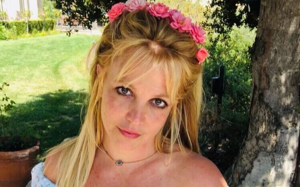 Britney Spears sem maquiagem, com uma coroa de flores na cabeça em um gramado a céu aberto