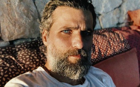 Barbudo e de camiseta branca, Bruno Gagliasso faz cara séria em selfie; ao fundo, temos uma parede de pedras e um sofá alaranjado