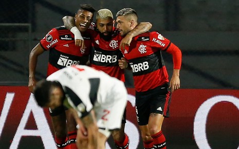 Bruno Henrique, Gabigol e Arrascaeta usam a camisa Rubro-Negra do Flamengo e se abraçam no estádio Defensores del Chaco, em Assunção, no Paraguai. Um jogador do Olimpía está abaixado e aparece na imagem.