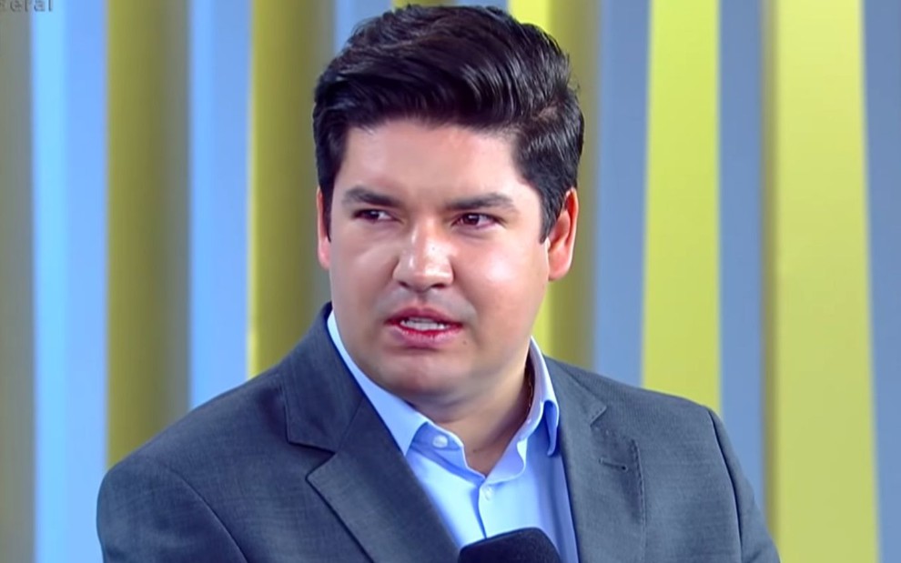 Bruno Peruka com terno e camisa azuis, olhando para o lado, no cenário de um telejornal