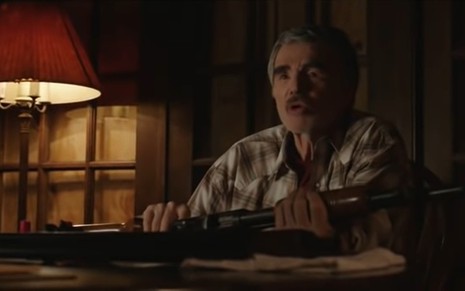 Burt Reynolds sentado, veste uma camisa xadrez, ao seu lado tem um abajur, e ele segura uma espingarda