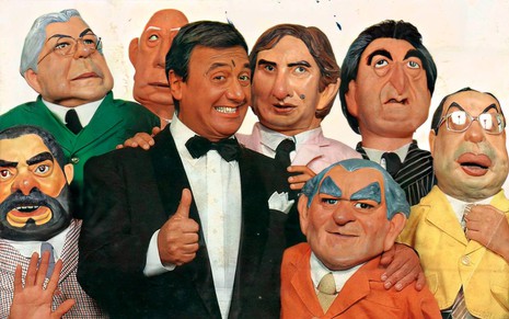 Agildo Ribeiro de smoking, fazendo sinal de joia e sorrindo, rodeado por seus bonecos que imitavam políticos
