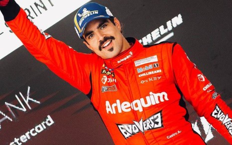 Caio Castro com seu uniforme vermelho de piloto de corrida, com um braço levantado