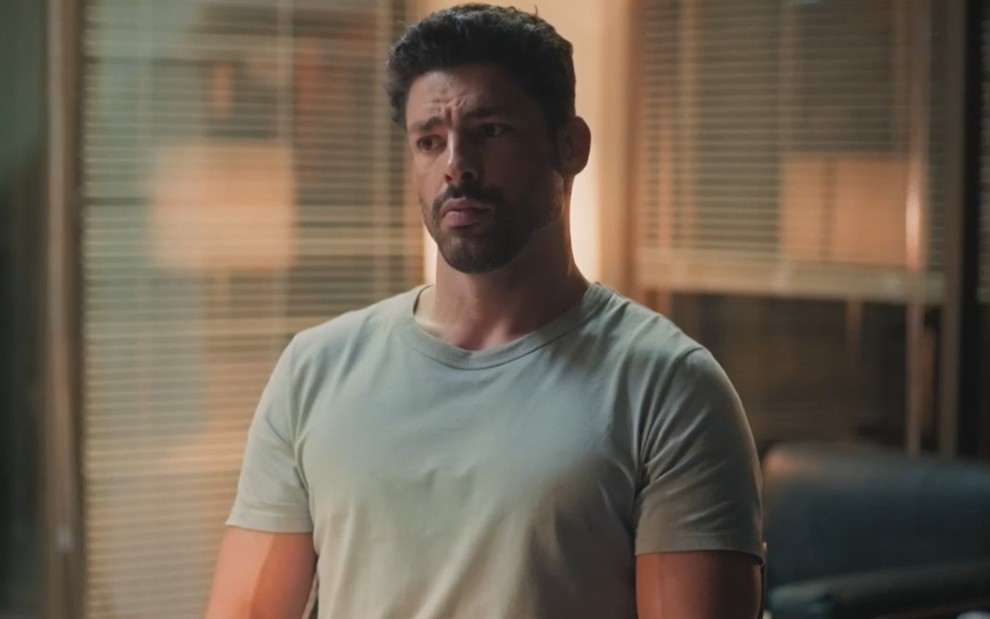 O ator Cauã Reymond em cena como Caio em Terra e Paixão, com expressão séria, camiseta cinza de manga curta, em escritório