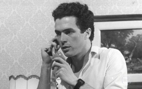O ator Caíque Ferreira segurando telefone, em cena de novela, em foto em preto e branco