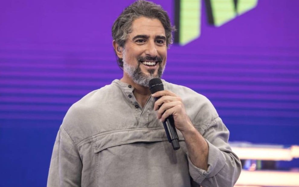 Marcos Mion na apresentação do Caldeirão: ele está com blusa cinza e segura microfone de mão