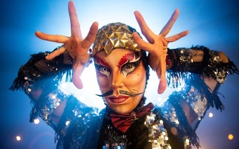 O drag king Don Valentim está maquiado, com as mãos na altura da cabeça e faz um movimento com os dedos