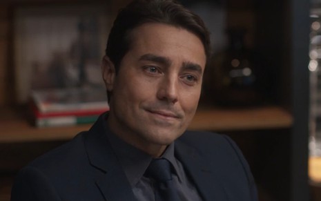 O ator Ricardo Pereira com leve sorriso, terno e gravata, em cena de Cara e Coragem