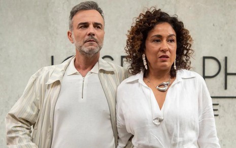 Os atores Marcelo Valle e Raquel Rocha lado a lado, com roupas brancas e expressões de preocupação