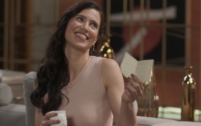 A atriz Mel Lisboa como Regina em Cara e Coragem; ela está sentada, olhando para cima e sorrindo, com um bilhete na mão