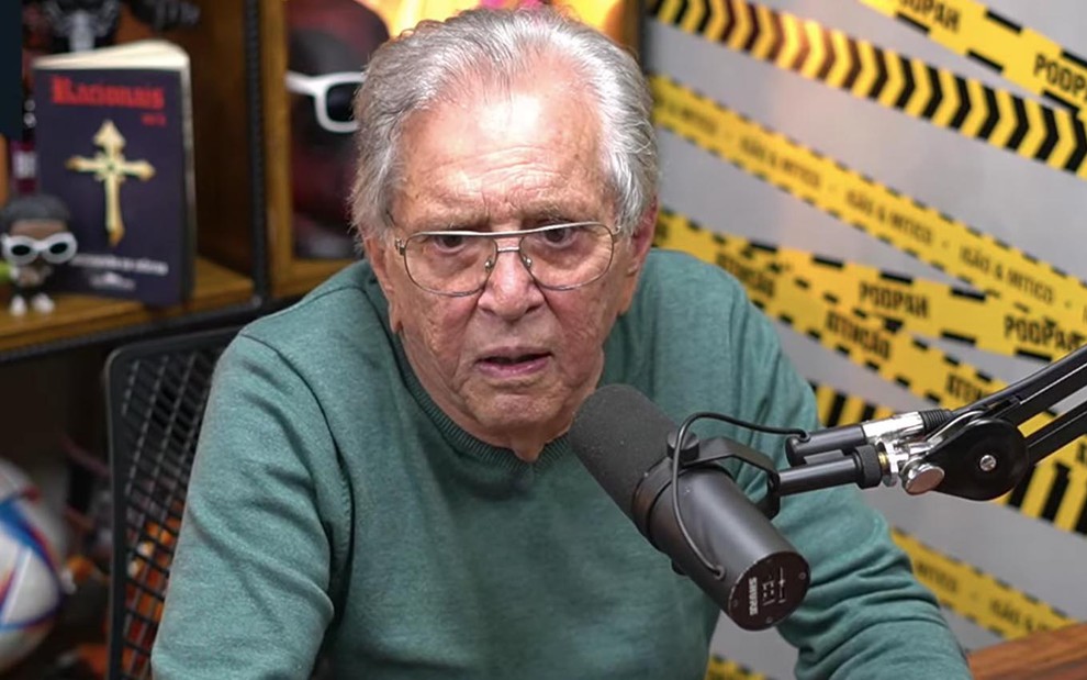 O comediante Carlos Alberto de Nóbrega com uma camisa verde de manga comprida em um estúdio de gravação em frente a um microfone