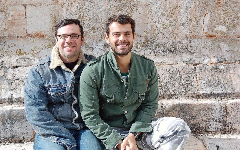João Emanuel Carneiro e Carmo Dalla Vecchia estão sentados, sorrindo e vestem roupas de frio