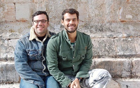 João Emanuel Carneiro e Carmo Dalla Vecchia estão sentados, sorrindo e vestem roupas de frio