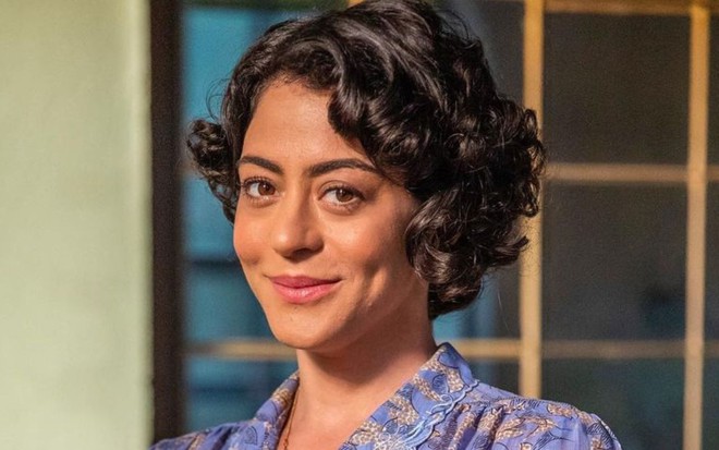 Carol Castro sorri discretamente e usa roupas de época em caracterização da novela Amor Perfeito