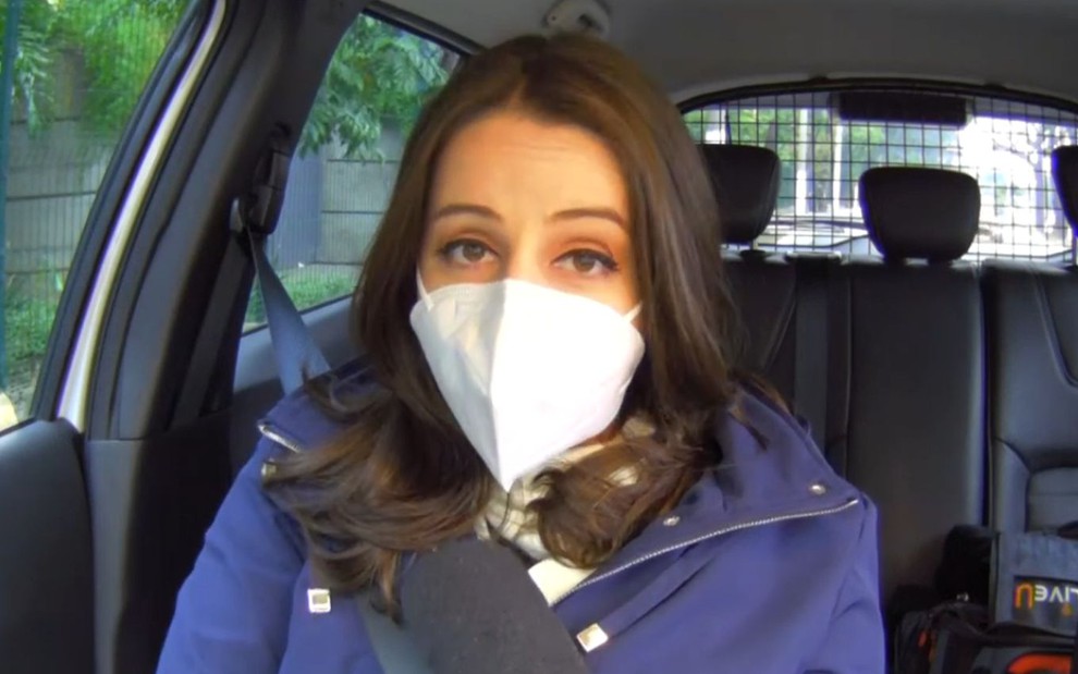 Carol Ianelli de máscara branca, casaco azul, sentada no banco da frente do carro da equipe da Globo