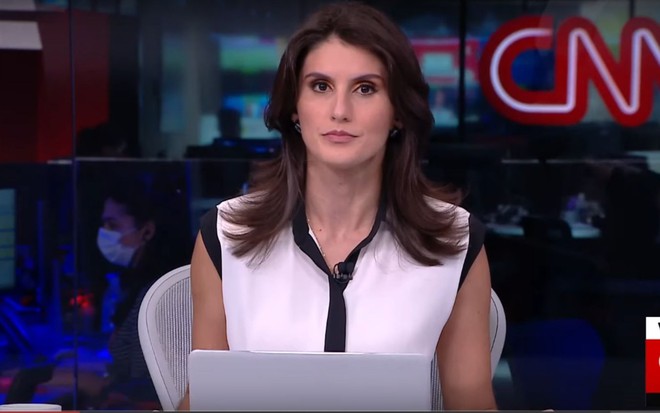 Carol Nogueira na bancada no Jornal da CNN. Ela está sentada, em frente a um notebook e veste uma camiseta branca. No fundo é possível ver o logo da CNN.