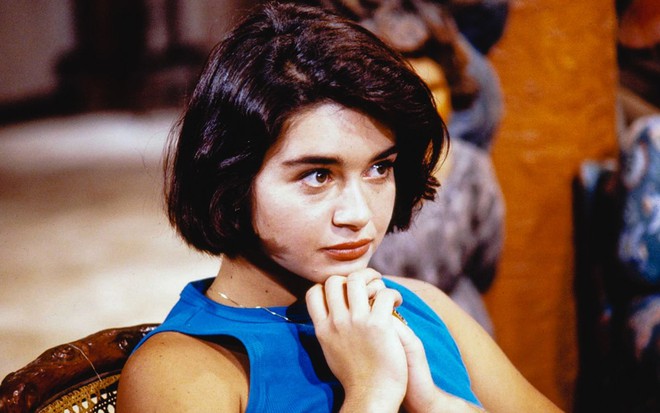 Em cena de Mulheres de Areia, Alexandra Marzo está sentada, olhando para alguém; ela usa blusa azul