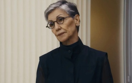 Cassia Kis com expressão séria, óculos e blusa preta de gola alta em cena como Cidália na novela Travessia