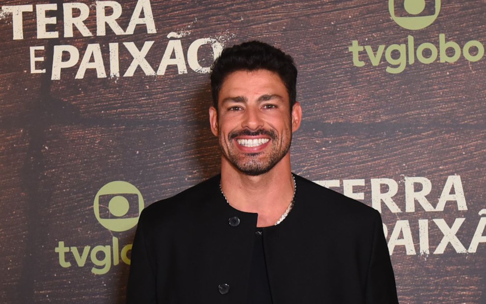 Cauã Reymond sorri com uma blusa preta em frente a um background em se lê Terra e Paixão