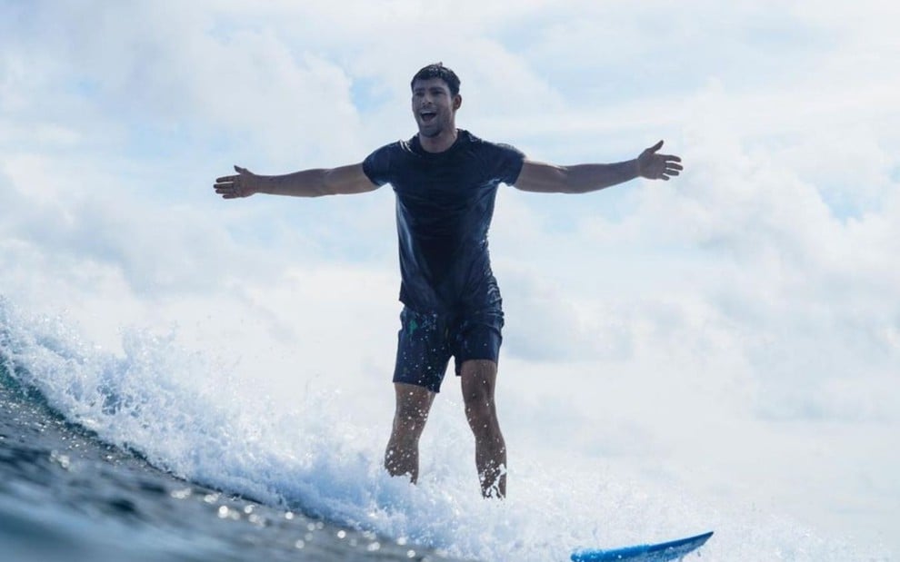 Cauã Reymond em cima de uma prancha pegando onda nas maldivas. Ele usa uma camiseta preta e um shorts preto e abre os braços posando para a câmera. No fundo é possível observar um céu nublado.