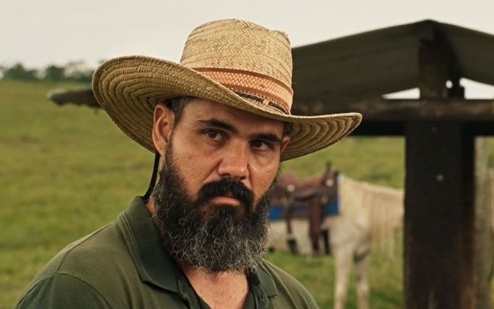 O ator Juliano Cazarré como Alcides em Pantanal; ele está de chapéu e olhando para o lado com cara séria em um pasto com gado atrás