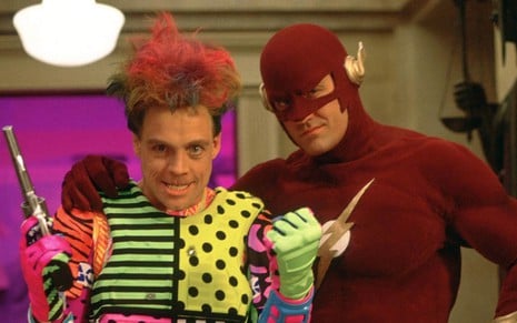 Mark Hamill tem visual bizarro e segura uma arma, enquanto John Wesley Shipp usa o traje do Flash; os dois posam abraçados para foto