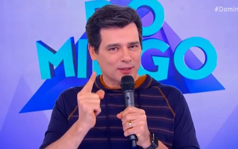 Celso Portiolli de blusa azul, no palco do Domingo Legal, com o dedo indicador apontado para frente e segurando microfone com a mão esquerda