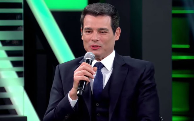 Celso Portiolli de terno e gravata escuros, segurando um microfone em um cenário de cores verde e preto