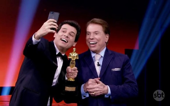 Celso Portiolli com o Troféu Imprensa em 2019, junto à Silvio Santos, sorrindo e tirando uma selfie. Celso está de preto e Silvio com um terno azul.