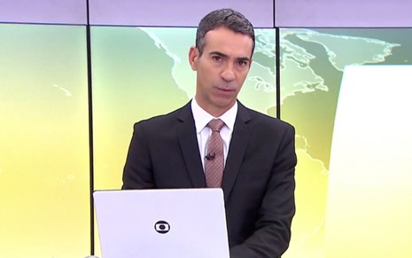 César Tralli apresentando o Jornal Hoje na Globo; ele usa um notebook e um telão está atrás dele