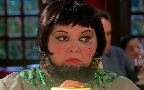 A atriz Fernanda Lobo como Mulher Barbada em Chocolate com Pimenta; ela está sentada a uma mesa, olhando ligeiramente para o lado com cara séria