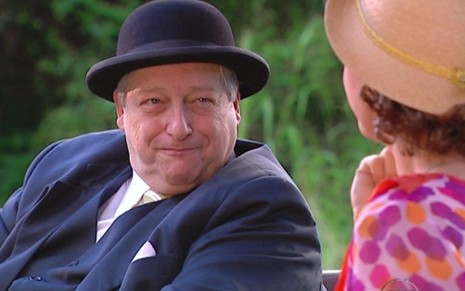 O ator Fúlvio Stefanini como Vivaldo em Chocolate com Pimenta; de chapéu, ele está sentado, olhando para o lado e quase rindo