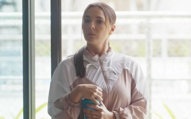 Em cena de Vai na Fé, Regiane Alves está com roupa esportiva rosa, em pé, com a expressão séria