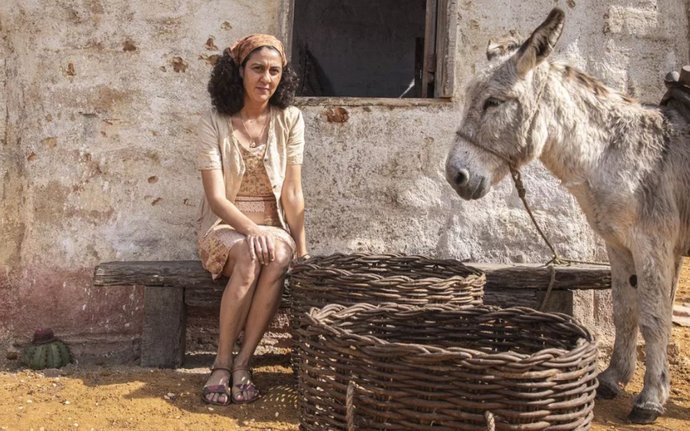 A atriz Clarissa Pinheiro como Tereza em Mar do Sertão; ela está sentada, com um lenço na cabeça, e as mãos nas pernas, ao lado de um burro e um balaio