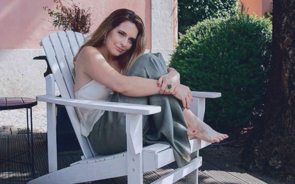 Cláudia Abreu posa sentada em uma cadeira e abraça as pernas