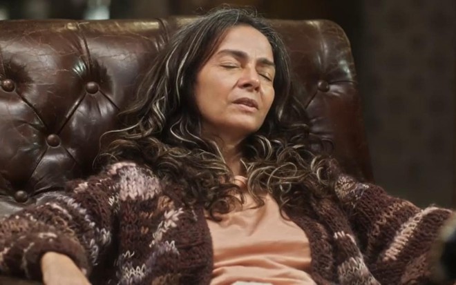 A atriz Claudia Ohana de olhos fechados, sentada em poltrona com expressão cansada, em cena de Vai na Fé