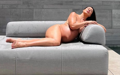 Grávida de sete meses Claudia Raia está nua, deitada de lado em um sofá cinza