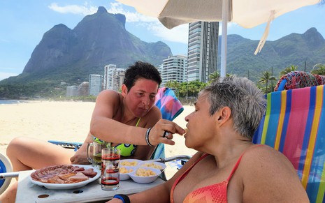 De biquíni em cadeira de praia, Adriane Bonatto dá petisco na boca da namorada em dia ensolarado