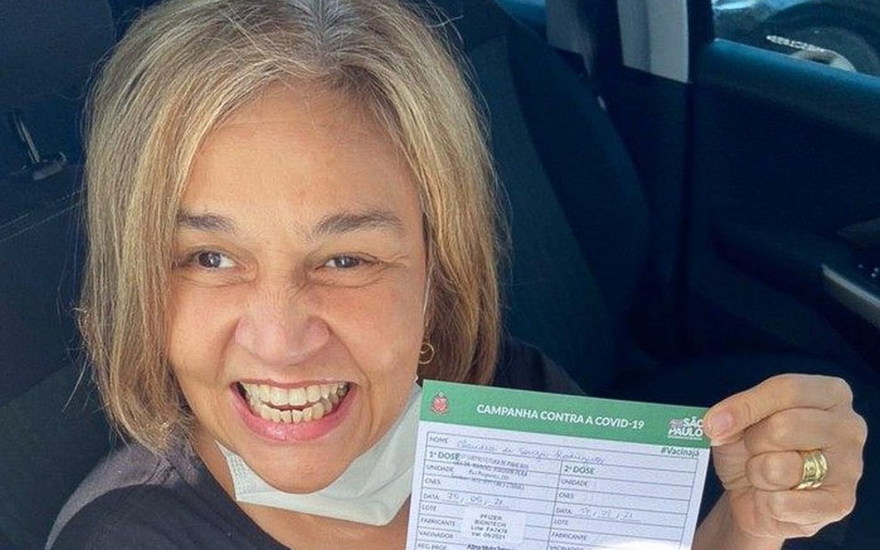 Claudia Rodrigues sorridente, cabelo chanel loiro, segurando cartão de vacinação
