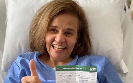 Claudia Rodrigues sorridente, de camisola azul, na cama de hospital, segurando o cartão de vacinação