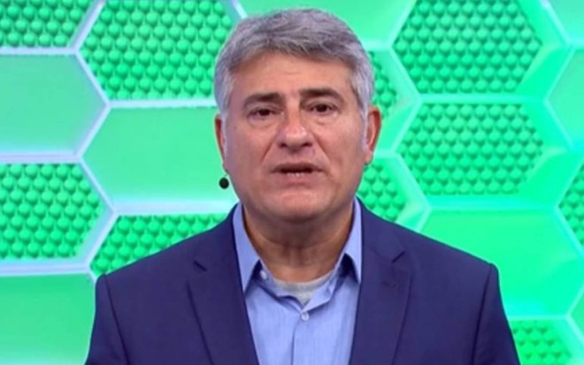 Cléber olha para frente, veste terno azul, camiseta azul e está na frente de uma parede verde