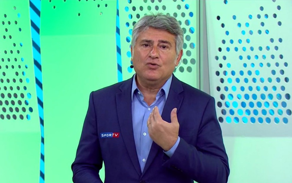 Cléber Machado usando um blazer azul marinho e uma camisa azul, com um fundo verde dos estúdios da Globo em São Paulo, durante uma transmissão